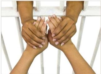 One pair of tanned hands holding another pair of tanned hands through white jail cell bars. 
